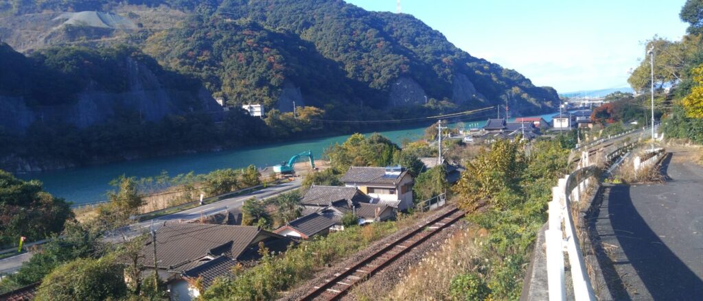 西部地域の高台から望む肥薩線線路と球磨川。線路と球磨川の間には家々、球磨川の背後には山と青空が広がっている